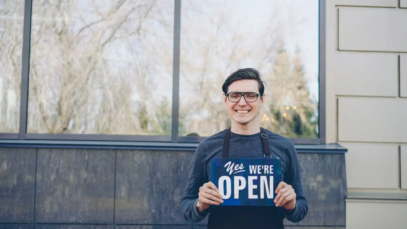 A smiling local small-business owner holding an "open" sign outside their shop